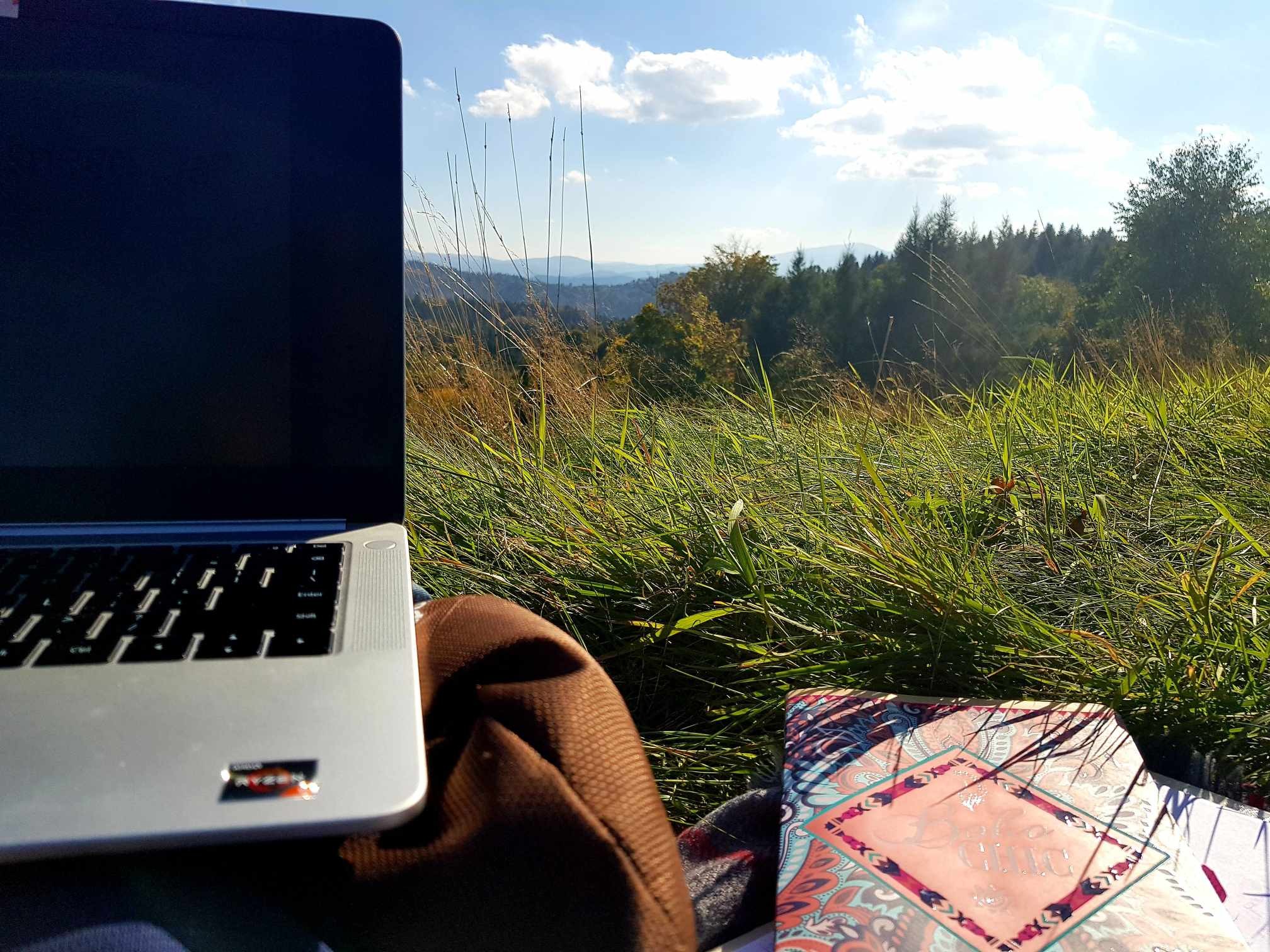 Praca w plenerze: rozłożony na kocu laptop, torba i notesy, w tle zielona trawa oraz góry w oddali.